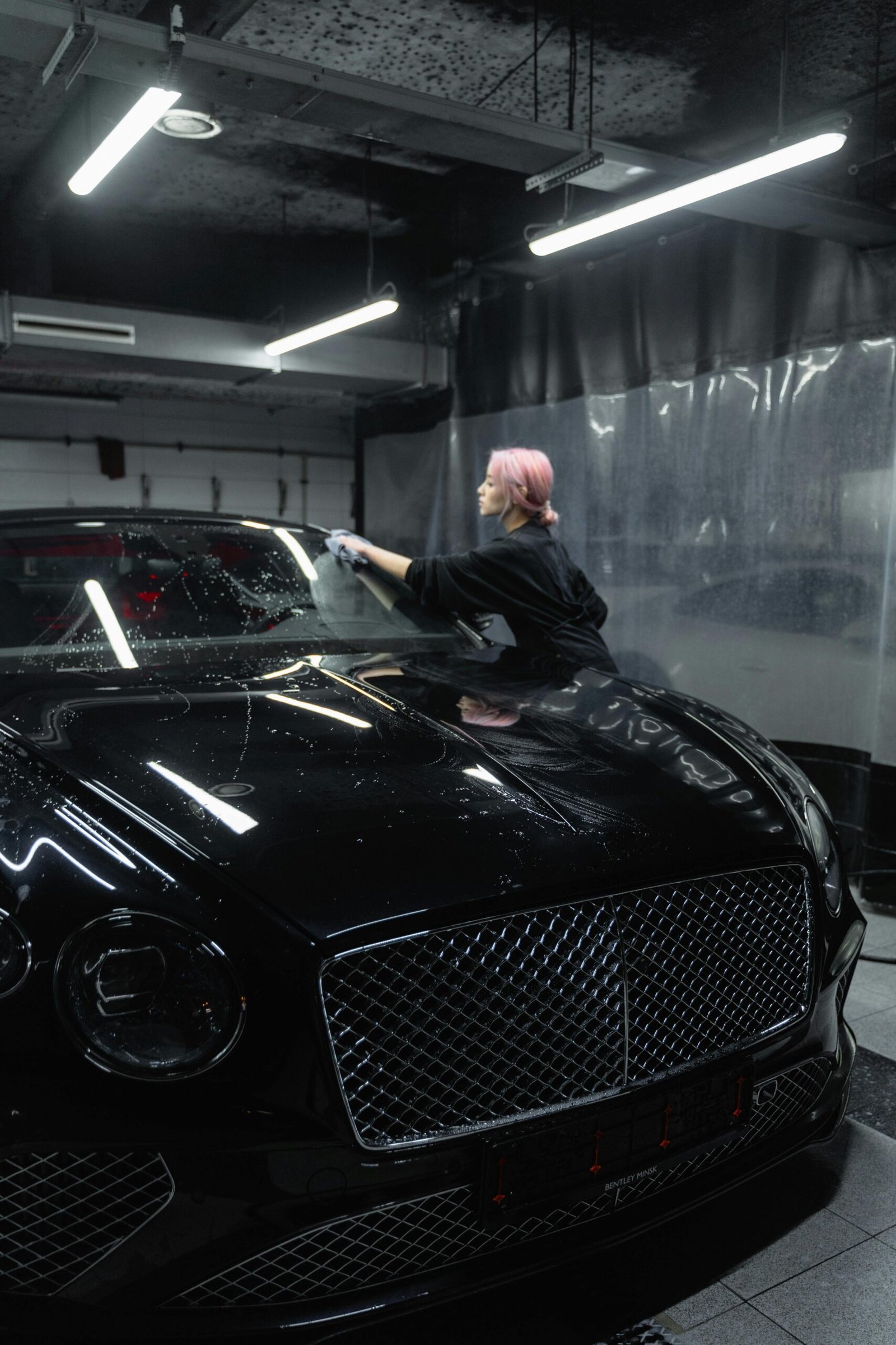 A woman cleans a sleek Bentley in an indoor car wash setting, adding shine.